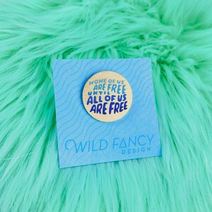 May include: A round, wooden pin with the text "NONE OF US ARE FREE UNTIL ALL OF US ARE FREE" in blue lettering. The pin is on a blue card with the words "WILD FANCY DESIGN" in a script font.