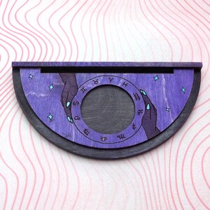 Puede incluir: Una rueda zodiacal de madera con una mancha p&uacute;rpura y un borde negro. La rueda presenta un c&iacute;rculo con los signos astrol&oacute;gicos y dos manos que se acercan la una a la otra.