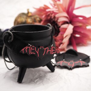 May include: A black cauldron with a bat-shaped necklace that says "They/Them" in red lettering. The necklace is hanging on the cauldron and there is a pink flower in the background.