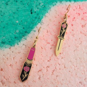 Op de afbeelding: Een paar gouden oorbellen met een roze lipstick en een zwart dolk ontwerp. Elke oorbel heeft een roze roos met een zwarte steel en bladeren.