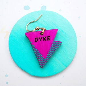 May include: Pink triangle-shaped wooden earring with a turquoise wave pattern and the word "DYKE" written in black lettering.