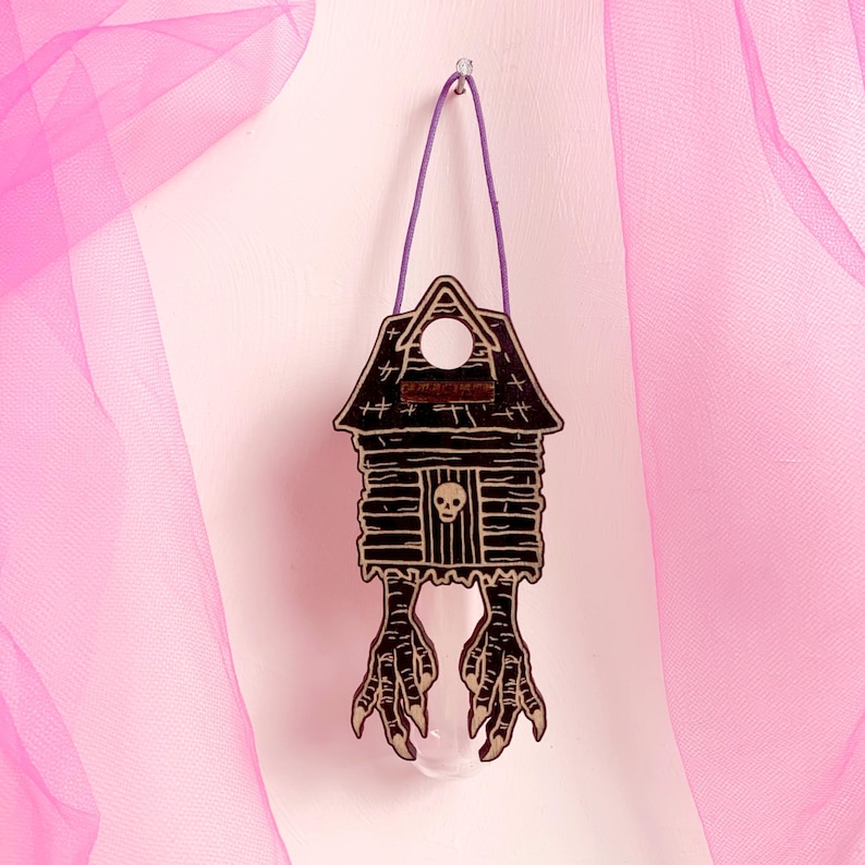 May include: A wooden haunted house ornament with a skull and bird feet design. The black and brown house has a purple string for hanging. The background is a light pink color.
