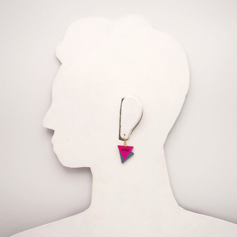 May include: Pink and blue triangle shaped earring with the word "DYKE" written on it.