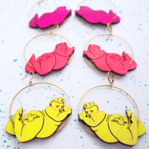 May include: Four pairs of hoop earrings with a gold-tone metal hoop and a wooden cutout of a person in a relaxed pose. Two pairs are pink and two pairs are yellow.