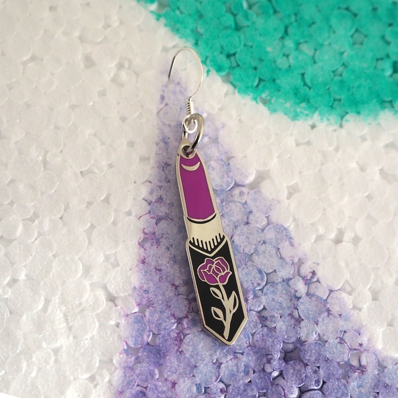 May include: A silver earring with a purple lipstick charm featuring a black rose design.