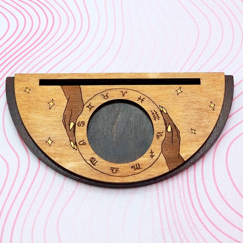 Puede incluir: Rueda zodiacal de madera con un centro marr&oacute;n oscuro y un anillo exterior marr&oacute;n claro. El anillo exterior tiene los signos del zod&iacute;aco grabados. Dos manos se acercan al centro de la rueda.