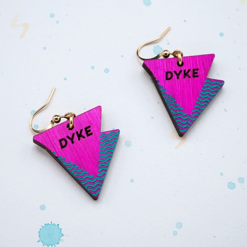 May include: A pair of pink triangle earrings with a teal wave pattern and the word "DYKE" written in black.