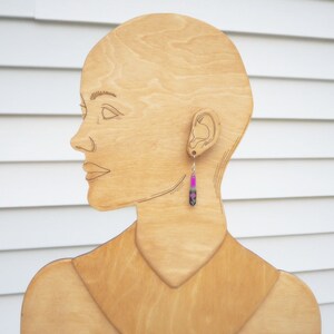May include: A wooden mannequin head with a pink and black dangle earring. The earring is made of a long, thin, rectangular piece of metal with a pink and black design.