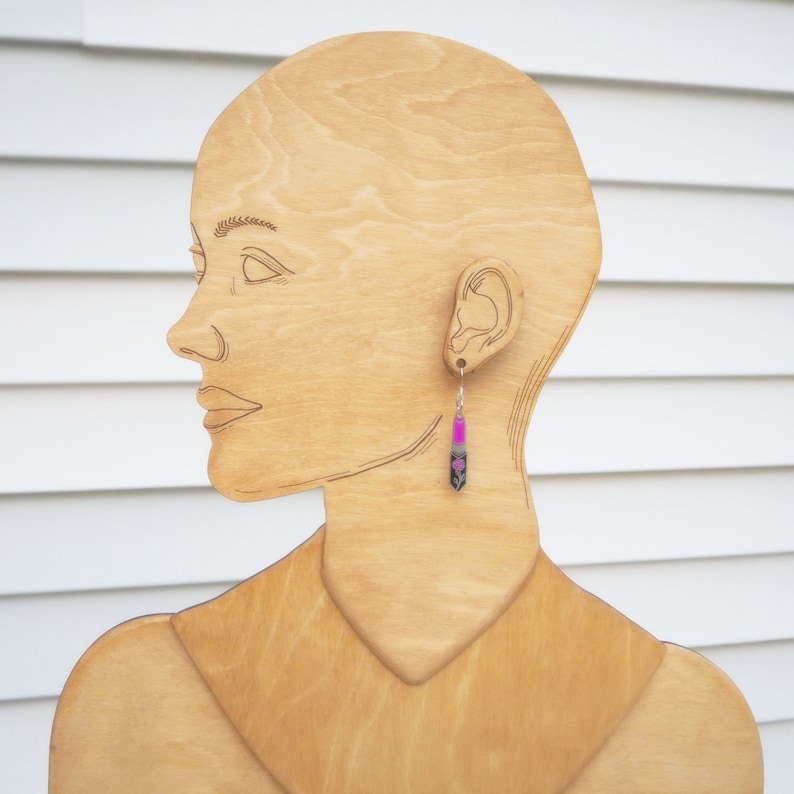 May include: A wooden mannequin head with a pink and black dangle earring. The earring is made of a long, thin, rectangular piece of metal with a pink and black design.