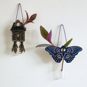 May include: Two wooden wall vases with glass tubes, each suspended by a purple cord. One is a house-shaped design with a skull and bird feet, the other is a blue moth with eye and moon details. Both hold greenery.