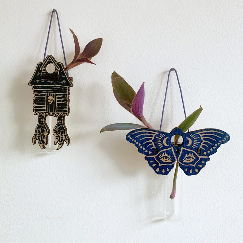May include: Two wooden wall vases with glass tubes, each suspended by a purple cord. One is a house-shaped design with a skull and bird feet, the other is a blue moth with eye and moon details. Both hold greenery.