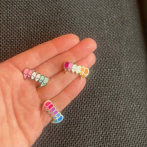 May include: Three enamel pins in the shape of a row of hearts. The first pin is pink, white, and light blue. The second pin is pink, purple, blue, and light blue. The third pin is orange, yellow, green, and blue.
