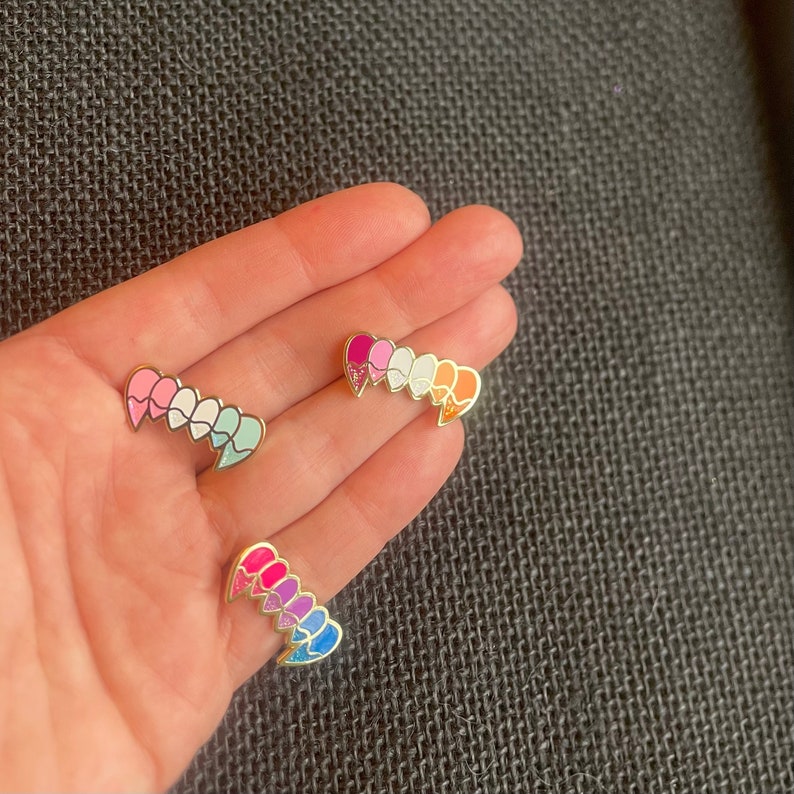 May include: Three enamel pins in the shape of a row of hearts. The first pin is pink, white, and light blue. The second pin is pink, purple, blue, and light blue. The third pin is orange, yellow, green, and blue.