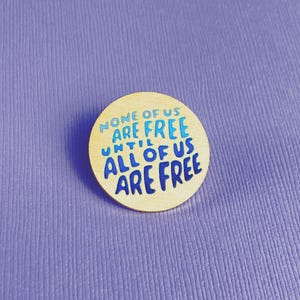 May include: A round wooden button with the text "NONE OF US ARE FREE UNTIL ALL OF US ARE FREE" in blue lettering. The button is set against a textured purple background.