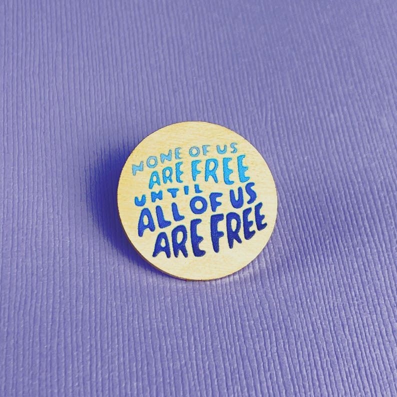 May include: A round wooden button with the text "NONE OF US ARE FREE UNTIL ALL OF US ARE FREE" in blue lettering. The button is set against a textured purple background.