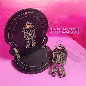 May include: A black wooden altar shelf and matching charm. The shelf is skull-shaped with a house design and the text "ALTAR SHELF ALSO AVAILABLE". The charm is a house with bird feet and a skull.