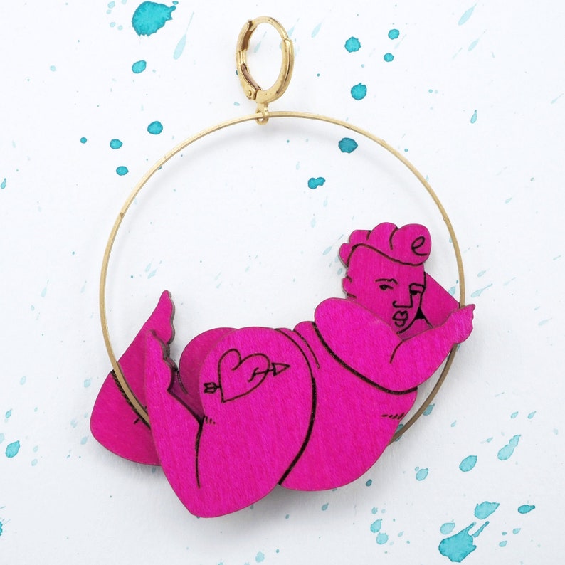 May include: A pink wooden earring featuring a figure of a person in a relaxed pose. The earring is suspended from a gold hoop.