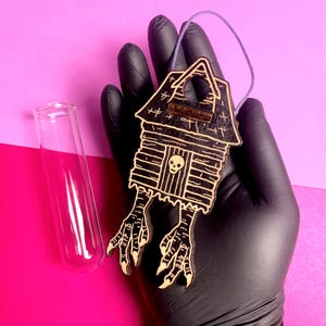 May include: A wooden, black and tan haunted house ornament with bird feet, a skull, and a purple string for hanging. A clear glass test tube is to the left of the ornament. The background is pink and purple.