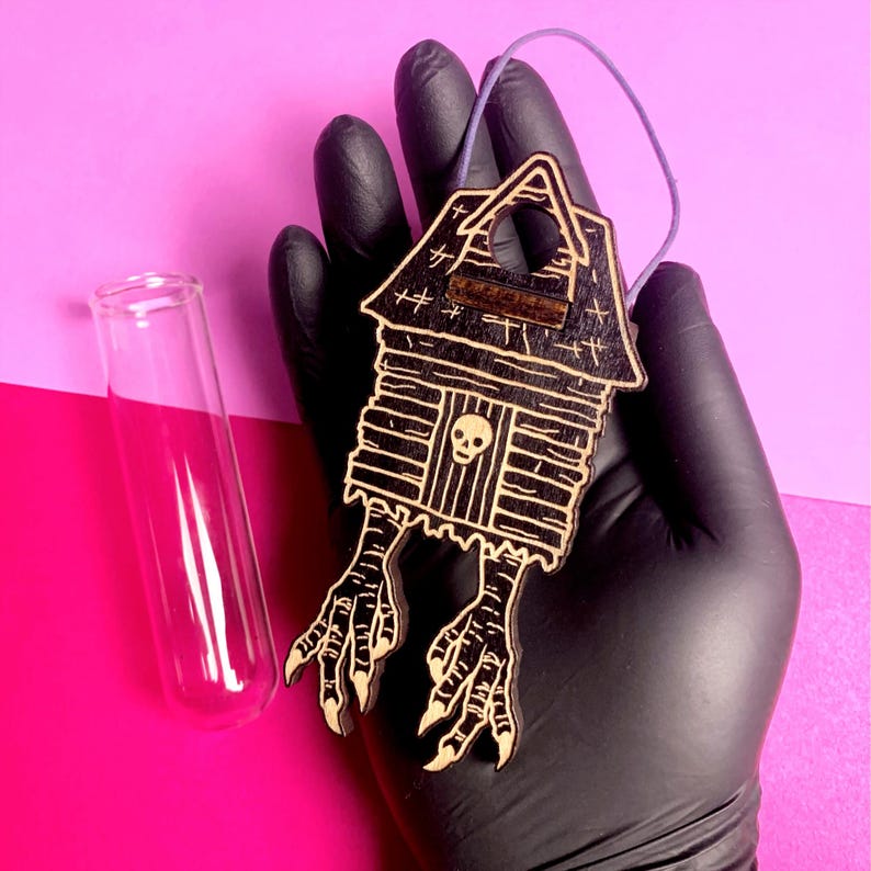 May include: A wooden, black and tan haunted house ornament with bird feet, a skull, and a purple string for hanging. A clear glass test tube is to the left of the ornament. The background is pink and purple.