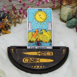 Puede incluir: Un soporte de madera para cartas de tarot con forma de media luna. El soporte tiene dos ranuras para las cartas y presenta un dise&ntilde;o decorativo de dos dagas y un patr&oacute;n floral. El soporte muestra una carta de tarot con la imagen de la luna y el texto "The Moon."
