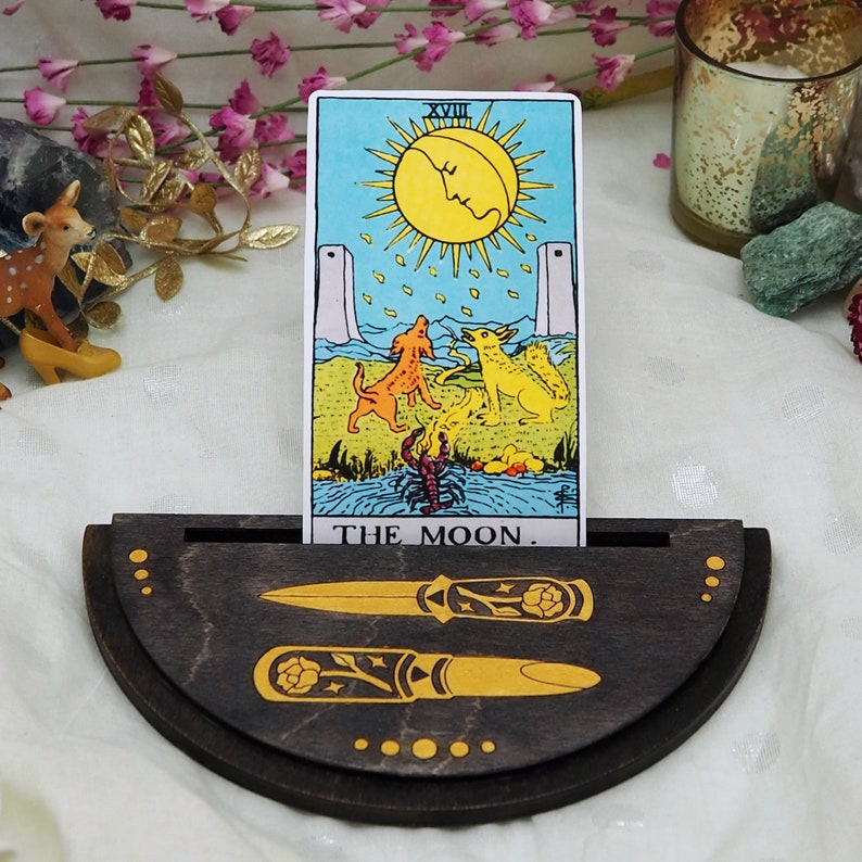 Puede incluir: Un soporte de madera para cartas de tarot con forma de media luna. El soporte tiene dos ranuras para las cartas y presenta un dise&ntilde;o decorativo de dos dagas y un patr&oacute;n floral. El soporte muestra una carta de tarot con la imagen de la luna y el texto "The Moon."