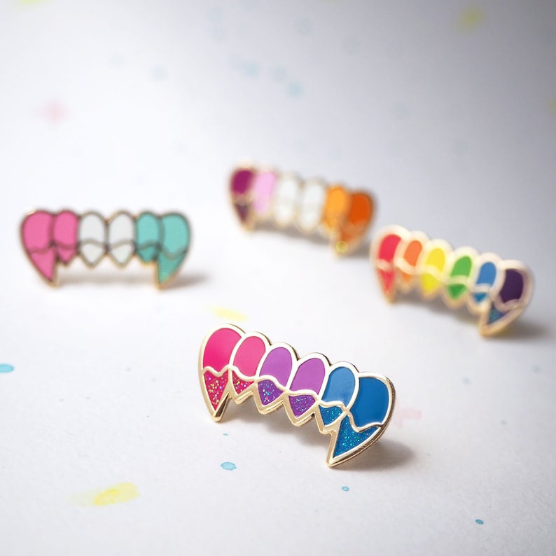 May include: A set of five enamel pins shaped like hearts, each with a different color of the rainbow. The pins are arranged in a line, with the red heart on the left and the purple heart on the right. The pins are gold-toned and have a sparkly finish.