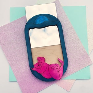 Puede incluir: Un espejo ovalado azul y blanco con una figura rosa de una persona en su interior. El espejo est&aacute; rodeado por un marco azul con un dise&ntilde;o de onda. La figura est&aacute; en una pose reclinada con un coraz&oacute;n en la cadera.