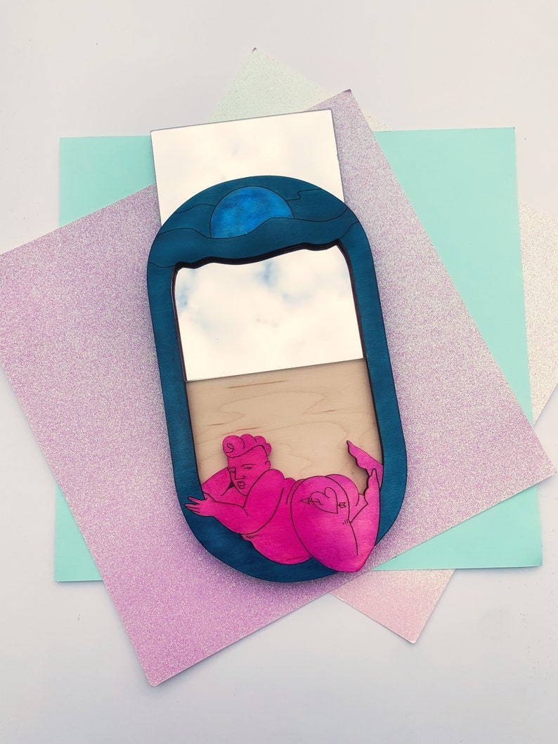 Puede incluir: Un espejo ovalado azul y blanco con una figura rosa de una persona en su interior. El espejo est&aacute; rodeado por un marco azul con un dise&ntilde;o de onda. La figura est&aacute; en una pose reclinada con un coraz&oacute;n en la cadera.