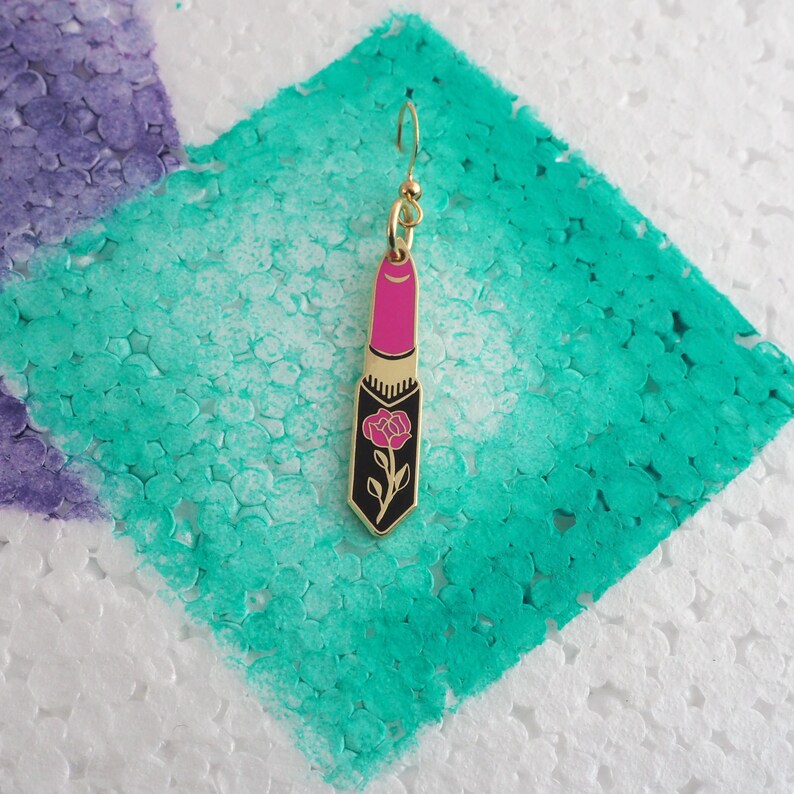 Op de afbeelding: Gouden oorbel met een roze lipstick-bedel met een zwart rozenmotief.