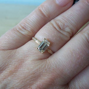 May include: A gold ring with an emerald cut gemstone. The ring is on a person's finger.