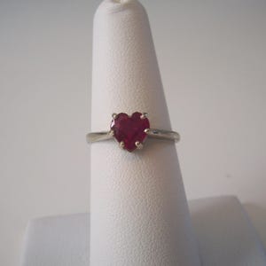 May include: A silver ring featuring a heart-shaped, deep red gemstone. The ring is displayed on a white finger form, showcasing the polished metal band and the vibrant color of the gemstone.
