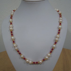 May include: A pearl necklace with alternating large, off-white pearls and small, red beads. The necklace has gold-colored accents between the pearls and beads. The necklace is displayed on a white mannequin.