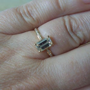 May include: A gold ring with a large emerald-cut diamond set in the center. The ring is on a person's finger.