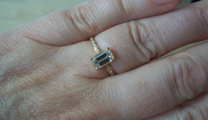 May include: A gold ring with a large emerald-cut diamond set in the center. The ring is on a person's finger.