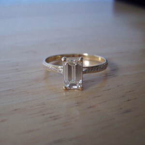 May include: A gold ring with a large, clear emerald-cut diamond set in the center. The band is accented with smaller diamonds.