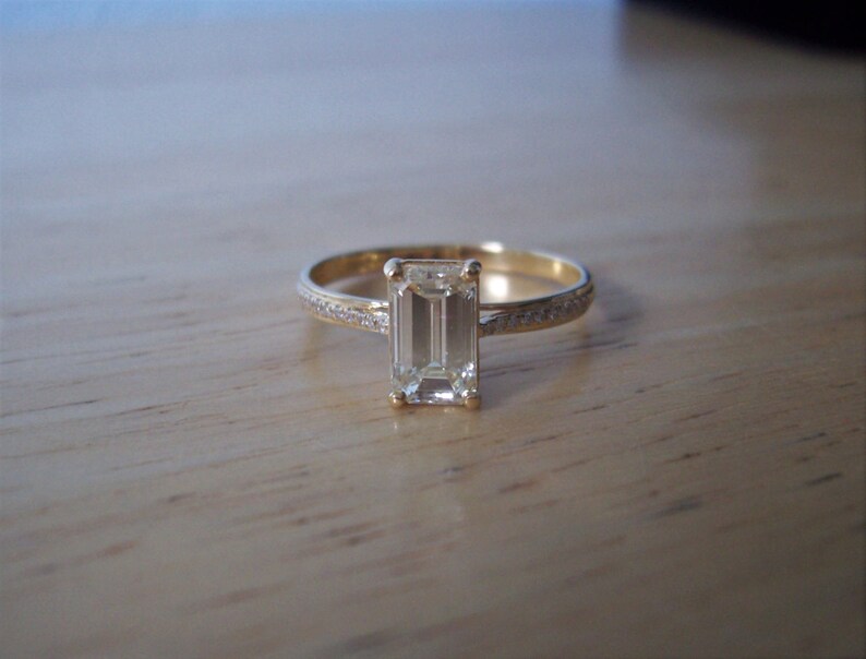 May include: A gold ring with a large, clear emerald-cut diamond set in the center. The band is accented with smaller diamonds.