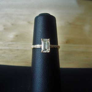May include: A gold ring with a clear emerald-cut diamond set in the center. The ring is displayed on a black velvet ring holder.