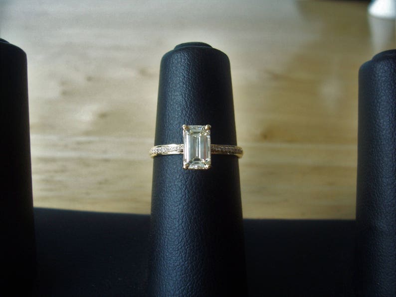May include: A gold ring with a clear emerald-cut diamond set in the center. The ring is displayed on a black velvet ring holder.