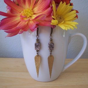 May include: A pair of gold earrings with a teardrop-shaped pendant and a square stone. The earrings are hanging from a white mug with pink and yellow flowers.