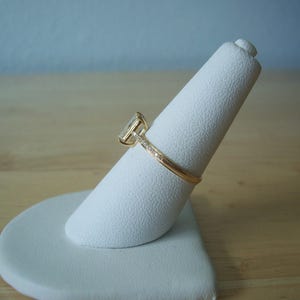May include: A gold ring with a square-shaped diamond set in a prong setting. The ring is on a white ring display stand.