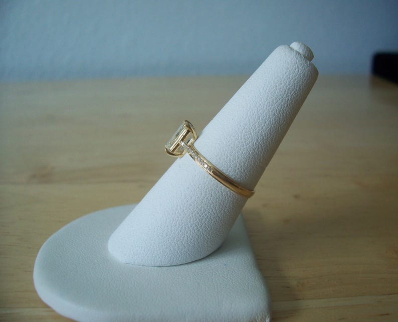 May include: A gold ring with a square-shaped diamond set in a prong setting. The ring is on a white ring display stand.