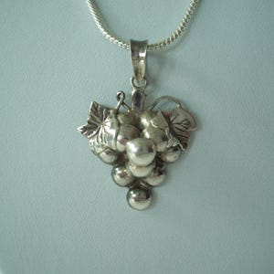May include: A silver pendant necklace featuring a cluster of grapes with leaves. The pendant is detailed with individual grape shapes and leaf veins. The necklace chain is a thin, silver snake chain. The pendant is approximately 1.5 inches in length.