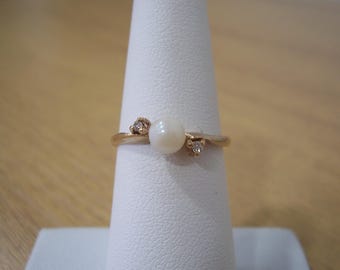 May include: A gold-toned ring featuring a central white pearl and two smaller accent stones. The ring is displayed on a white ring holder against a blurred wooden background. The design is delicate and elegant.