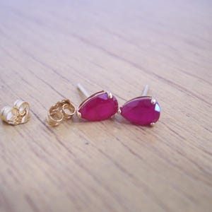 May include: A pair of teardrop-shaped stud earrings featuring vibrant pinkish-red gemstones set in gold-toned metal. The earrings include two matching gold-toned metal earring backs, all displayed together on a light-colored wooden surface.