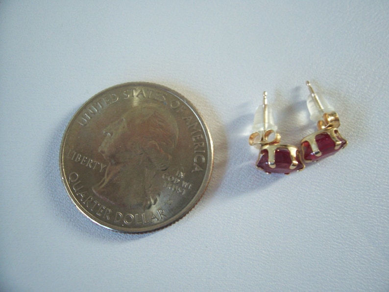 May include: A pair of gold stud earrings with red gemstone accents. The earrings are shown next to a US quarter for size comparison.