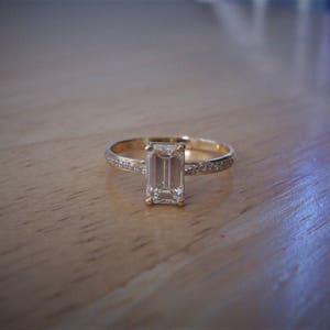 May include: A gold ring with a large, clear emerald-cut diamond set in the center. The band is thin and has small diamonds set around the center stone.
