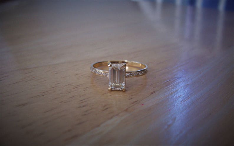 May include: A gold ring with a large, clear emerald-cut diamond set in the center. The band is thin and has small diamonds set around the center stone.