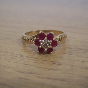 May include: A gold ring featuring a flower-shaped cluster of six round, deep red rubies surrounding a single central white diamond. The band of the ring displays an intricate, openwork scroll pattern on both sides of the central gemstone setting.