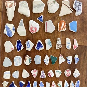 Peut inclure: Une collection de fragments de céramique de formes irrégulières, ornés de motifs et de couleurs variés, notamment bleu, rouge et marron. Les fragments semblent provenir de poteries cassées, disposés sur une surface en bois.