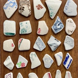 May include: A collection of twenty-five assorted ceramic pottery shards featuring various patterns and colors, including blue, red, green, and brown designs. Two silver-colored coins are positioned at the top, and a 15 cm ruler is at the bottom.